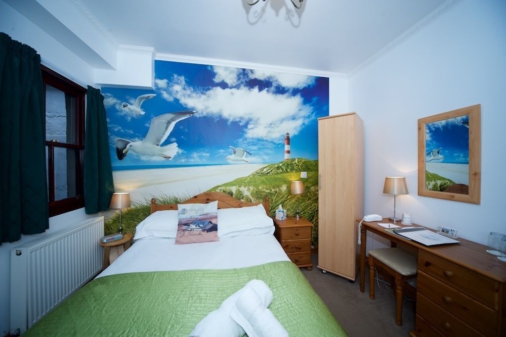 A bedroom with a large bed, a desk, a chair, a nightstand, a lamp, a mirror, and a painting of seagulls and a lighthouse.