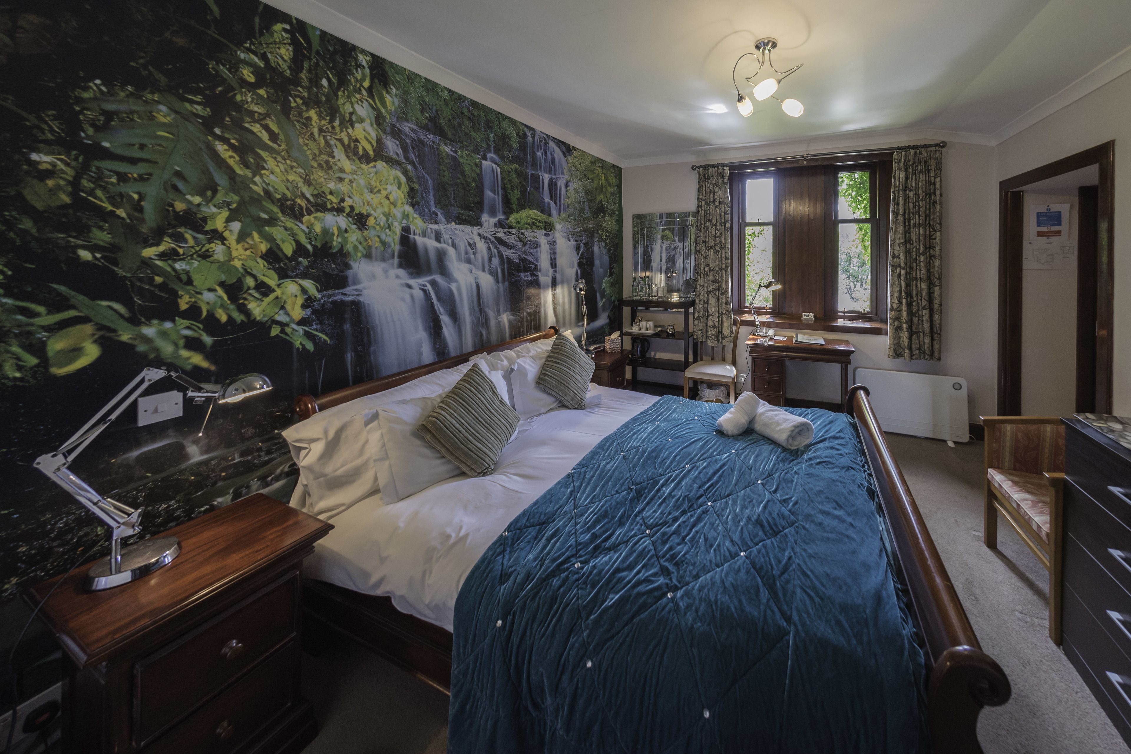 A bedroom with a large mural of a waterfall, a wooden bed with a blue quilt, a nightstand with a lamp, and a window with curtains.