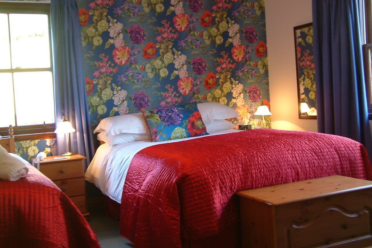 A bedroom with two beds, floral wallpaper, a wooden dresser, and a window with curtains.