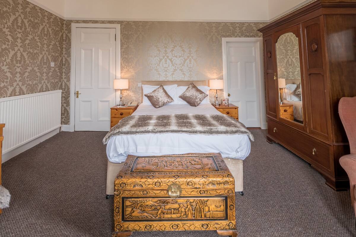 A bedroom with a large bed, two bedside tables with lamps, a wooden chest of drawers, and patterned wallpaper.