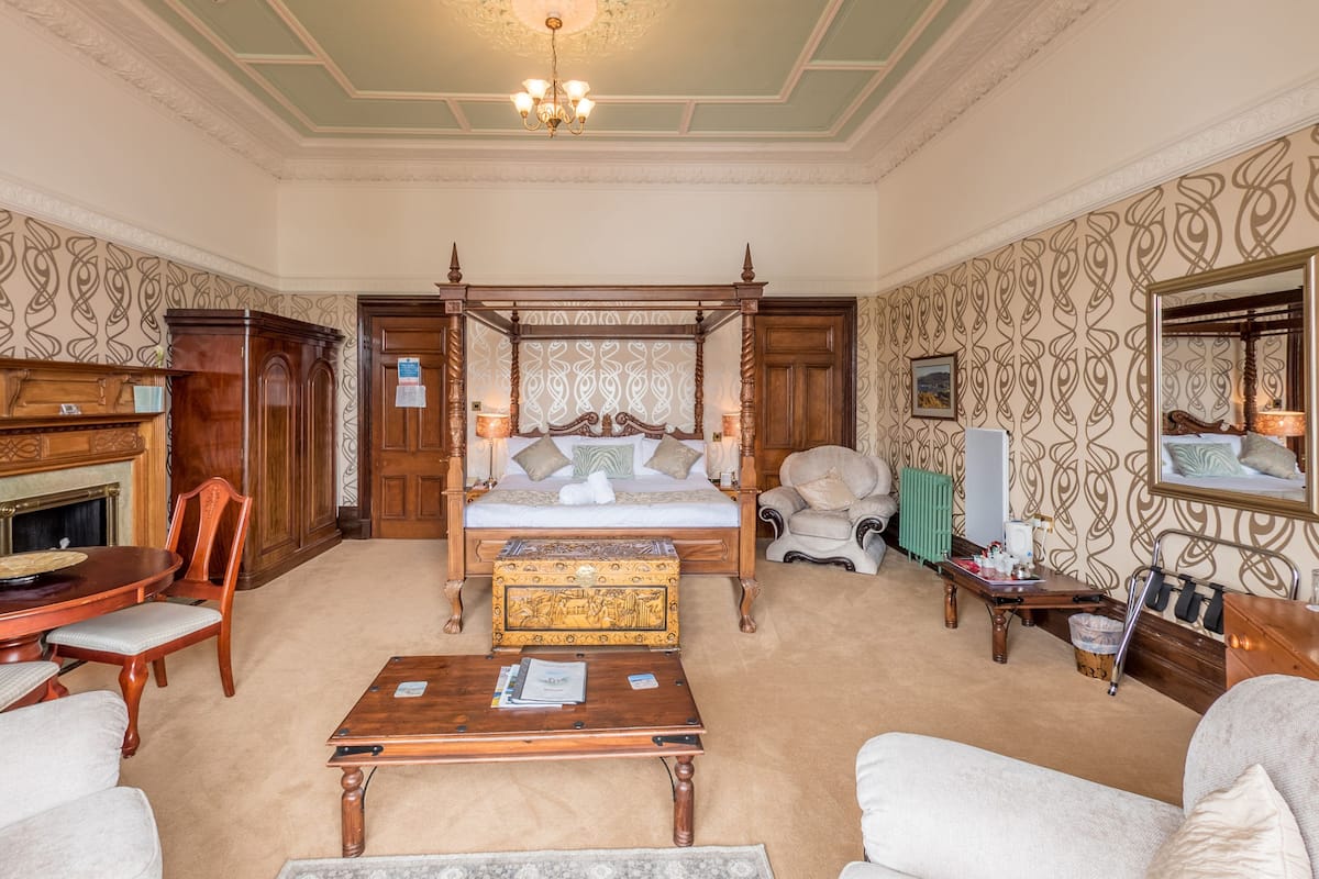 A traditional bedroom with a four-poster bed, a wooden dresser, a sitting area with a table and chairs, and a fireplace.