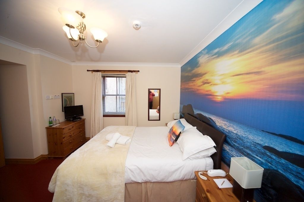 A hotel room with a large bed, a television on a wooden dresser, a desk with a telephone and a lamp, and a sunset mural on the wall.