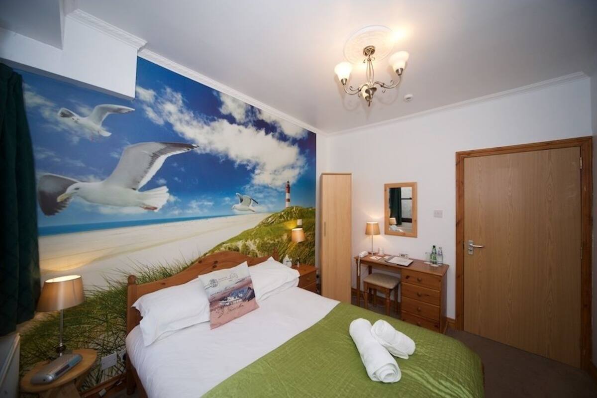 A hotel room with a mural of seagulls flying over a beach, a bed with a green bedspread, a desk with a chair, and a wooden door.