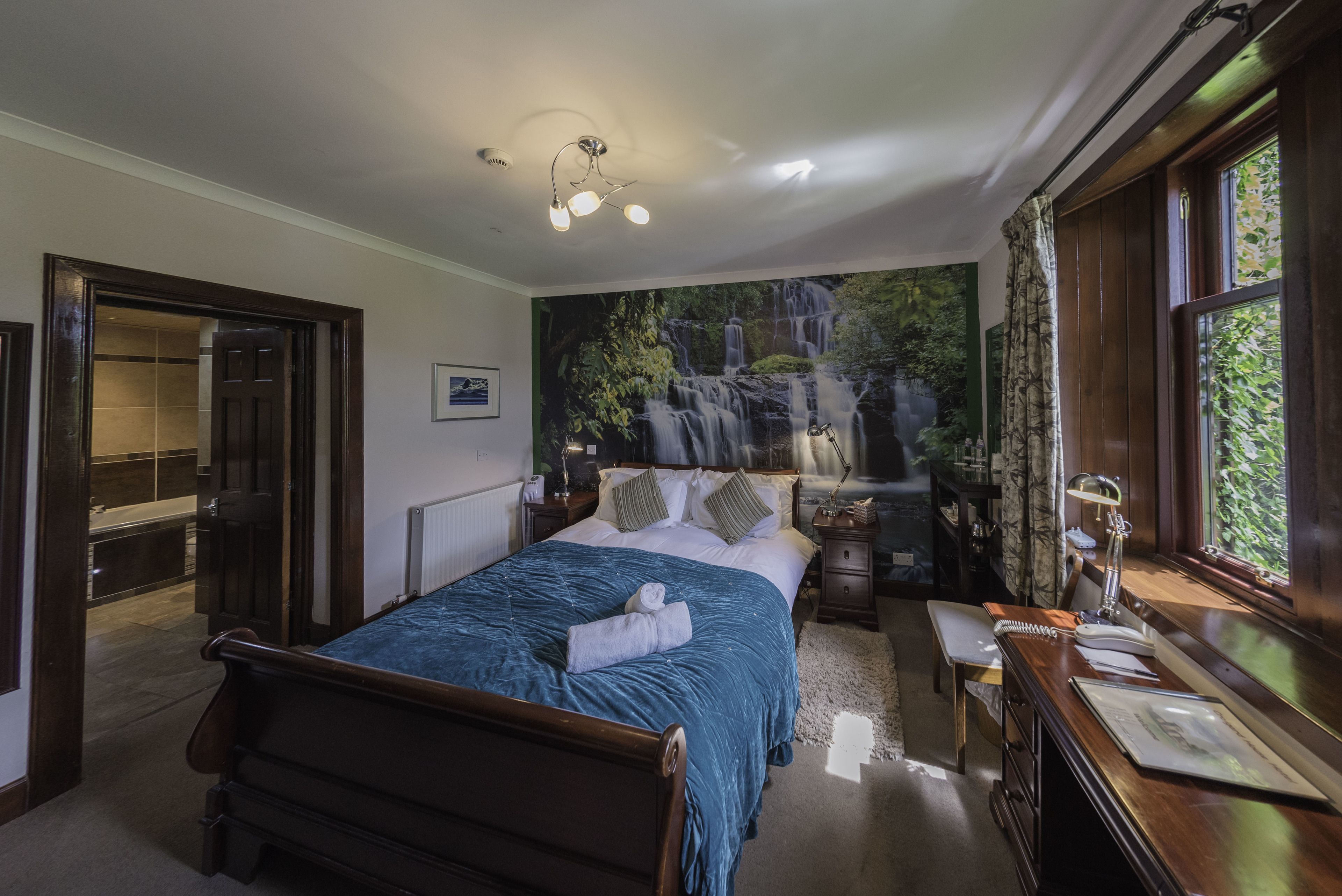 A bedroom with a large bed, a desk with a lamp, a window with curtains, a painting of a waterfall on the wall, and a door leading to another room.