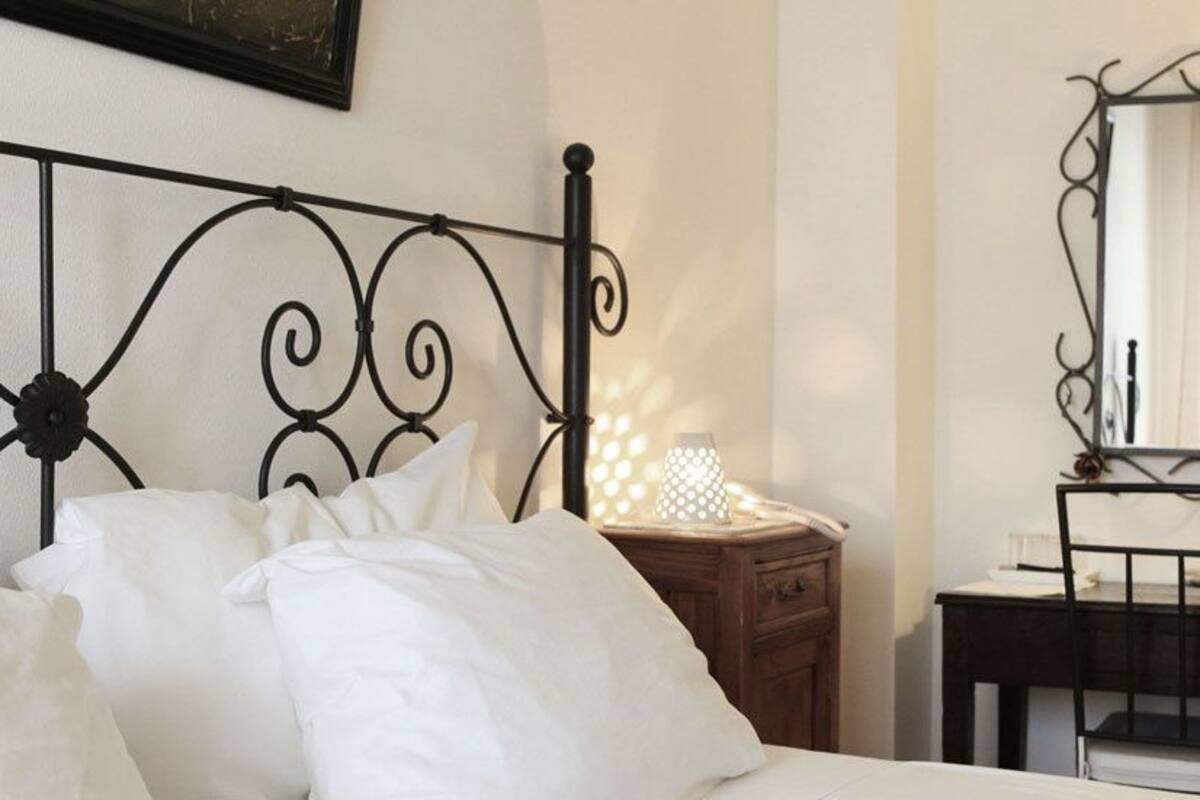 A neatly made bed with white linens, a bedside table with a lamp, a mirror, and a small wooden nightstand.