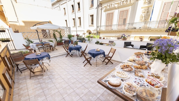 An outdoor patio with tables and chairs, a buffet table with food, and a building with balconies and decorative elements in the background.
