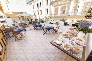 An outdoor patio with tables and chairs, a buffet table with food, and a building with balconies and decorative elements in the background. - Guesthouse B&B Garibaldi (Trapani)