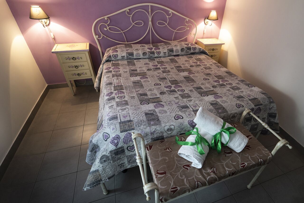 Un llit ben fet amb un edredó estampat, dues taules de nit amb làmpades i un banc amb una tovallola.