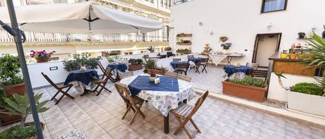 A rooftop terrace with tables and chairs, potted plants, and a white umbrella.