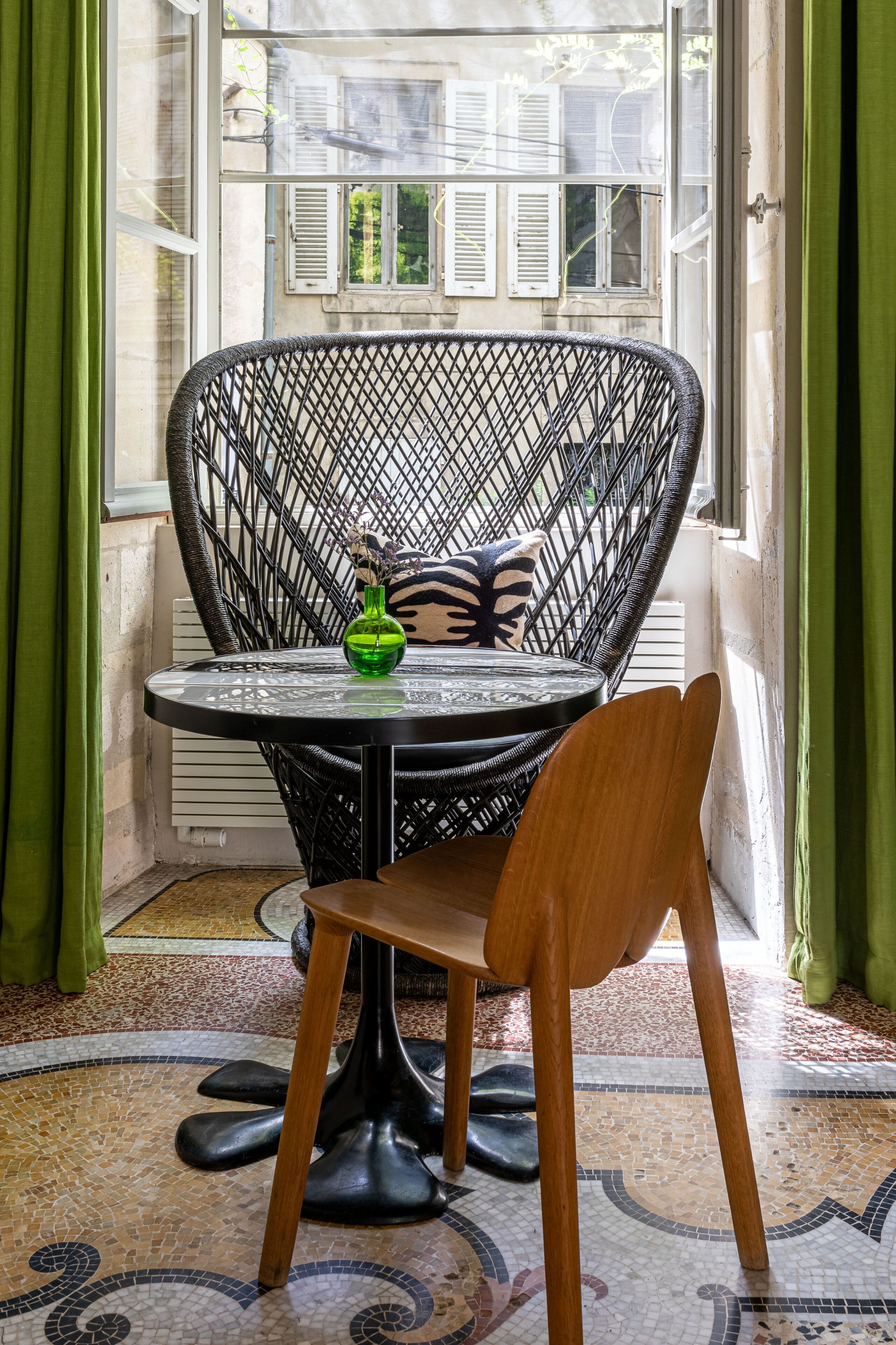 A room with a round table, a wicker chair, a green vase, and a patterned cushion.
