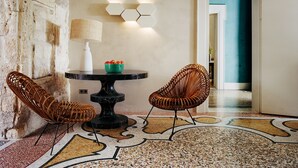 Een kamer met een ronde tafel, twee rieten stoelen, een schaal fruit, een lamp en een zeshoekige wandlamp.