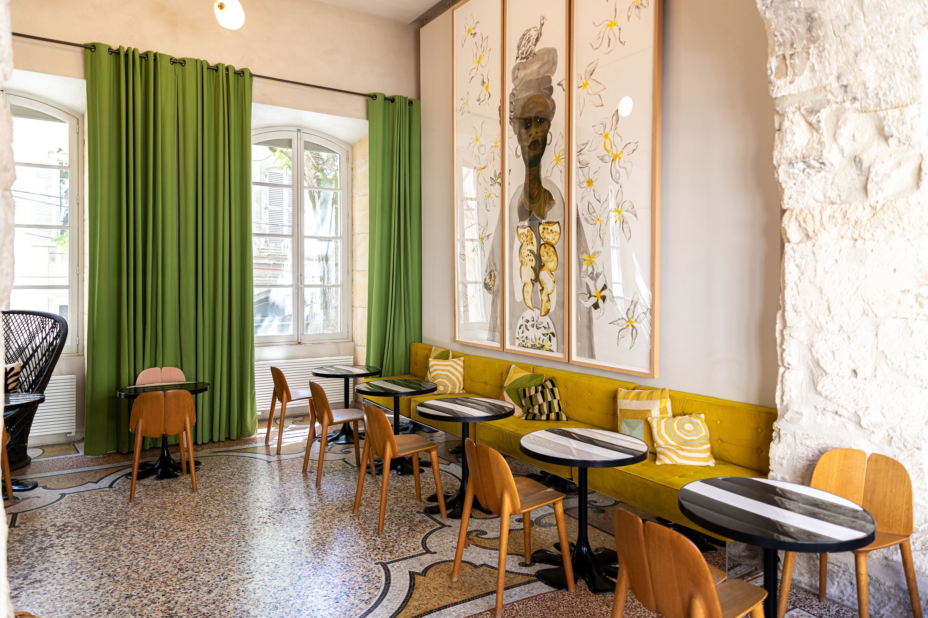 Une pièce avec un canapé vert, des chaises et des tables en bois, une fenêtre avec des rideaux verts et un mur orné d’œuvres d’art florales.