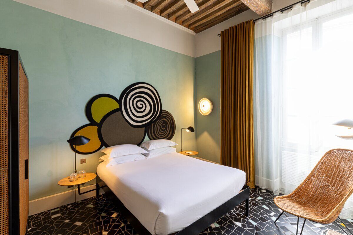 Une chambre d’hôtel avec un lit, une chaise, un tableau décoratif au mur, une table de chevet et une fenêtre avec des rideaux.