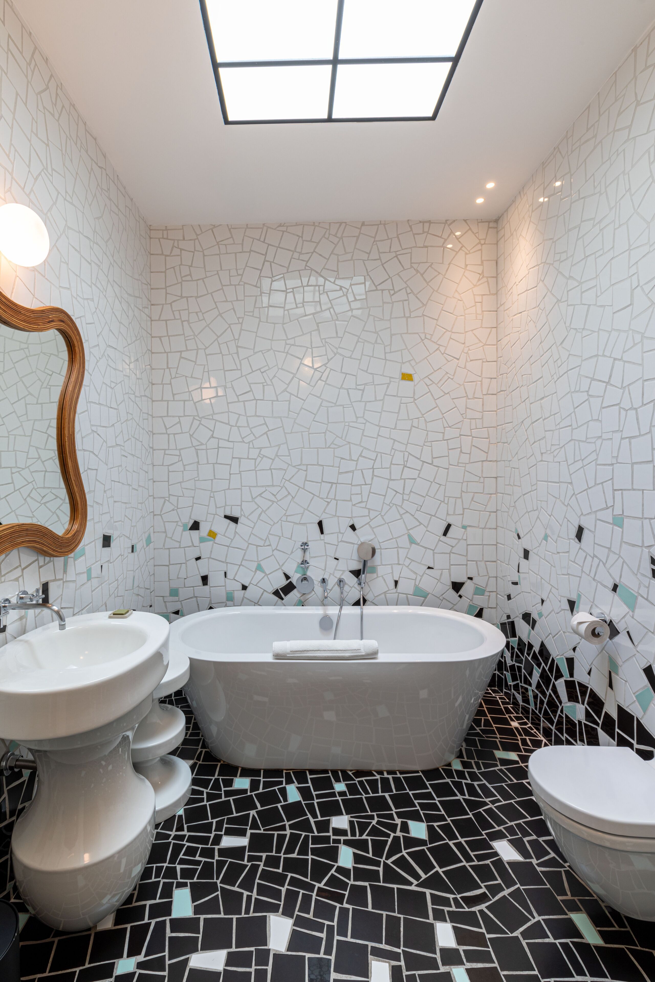 A bathroom with a freestanding bathtub, a sink, and a toilet. The walls are tiled with a mosaic pattern, and there is a skylight above.