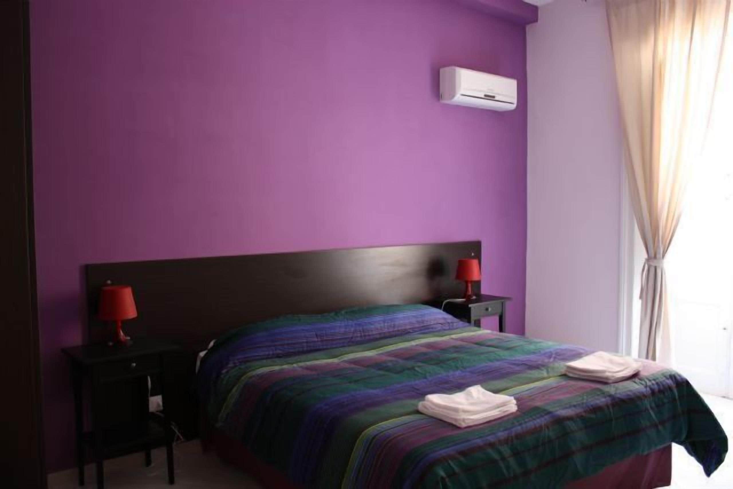 A bedroom with a purple wall, a bed with a multicolored blanket, two bedside tables with lamps, and an air conditioning unit.