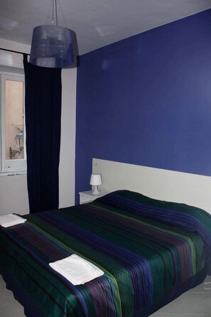 Quarto com cama, mesa de cabeceira, abajur e janela com cortinas.