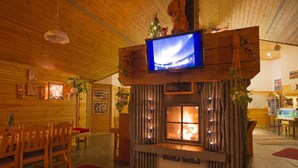 Interior acogedor de una cabaña con detalles de madera, una chimenea, un televisor montado en una viga de madera y una zona de comedor con muebles de madera.