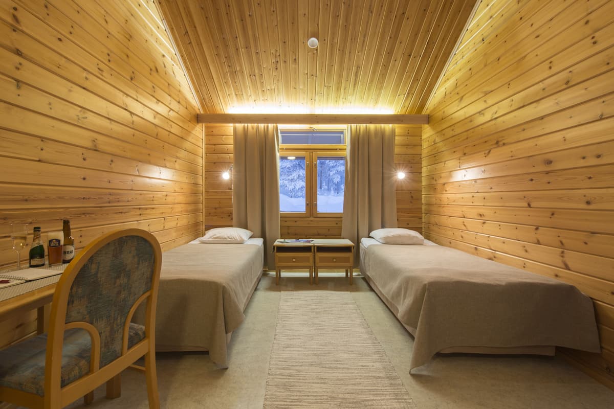 A cozy cabin room with two beds, a desk with a chair, and a window with a view of snow-covered trees.