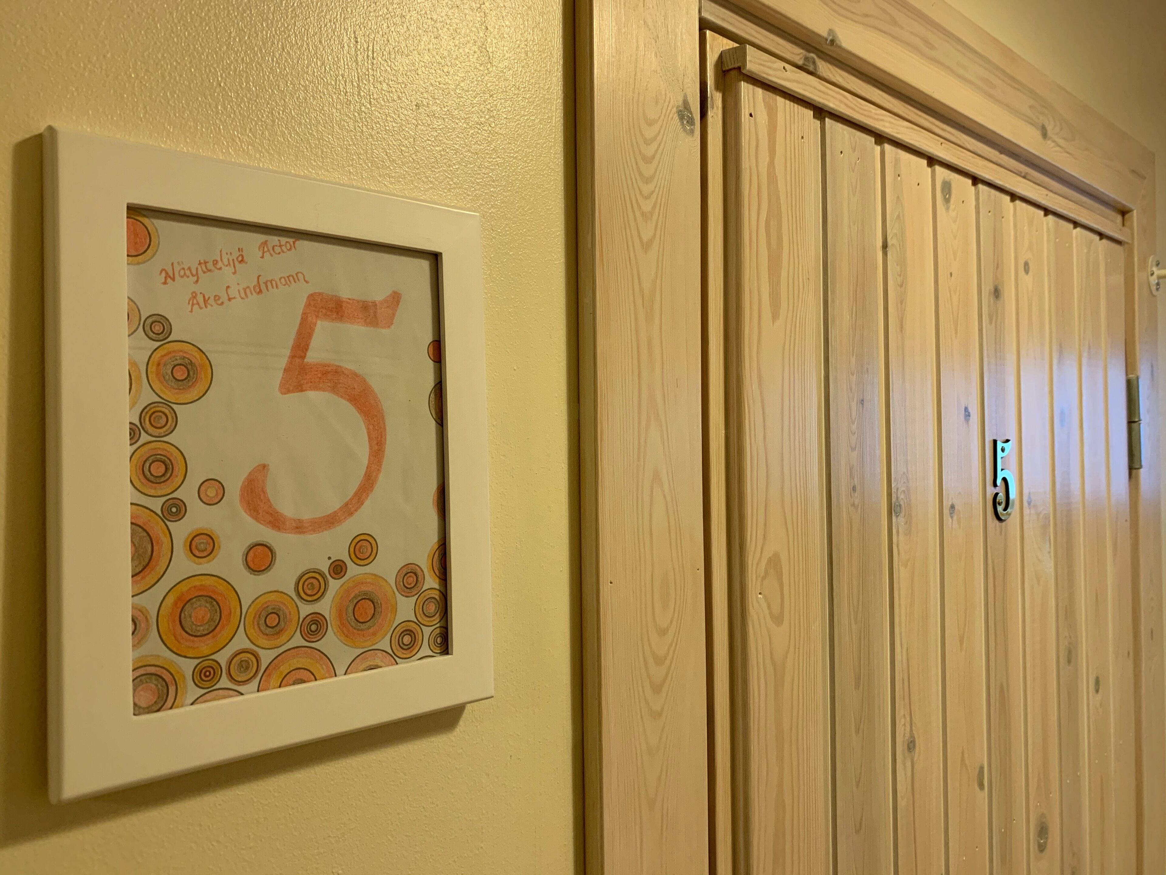 A framed artwork with the number 5 and text in a foreign language, a wooden door with a number 5 handle, and a wall with a textured finish.