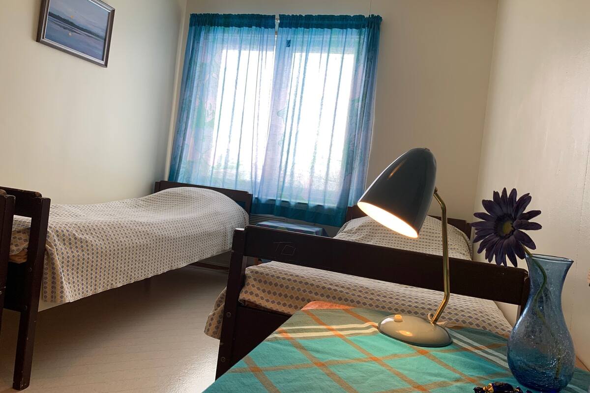 A room with two single beds, a desk with a lamp, a vase with a flower, and a window with blue curtains.