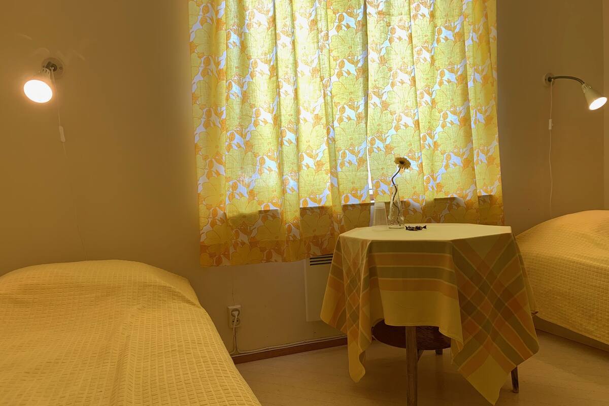 A room with two beds, a small table with a lamp, and a window with floral curtains.