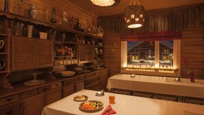 Interior acogedor de una cabaña de madera con una mesa de comedor preparada para dos comensales, una zona de cocina con armarios de madera y un ventanal con vistas al exterior.
