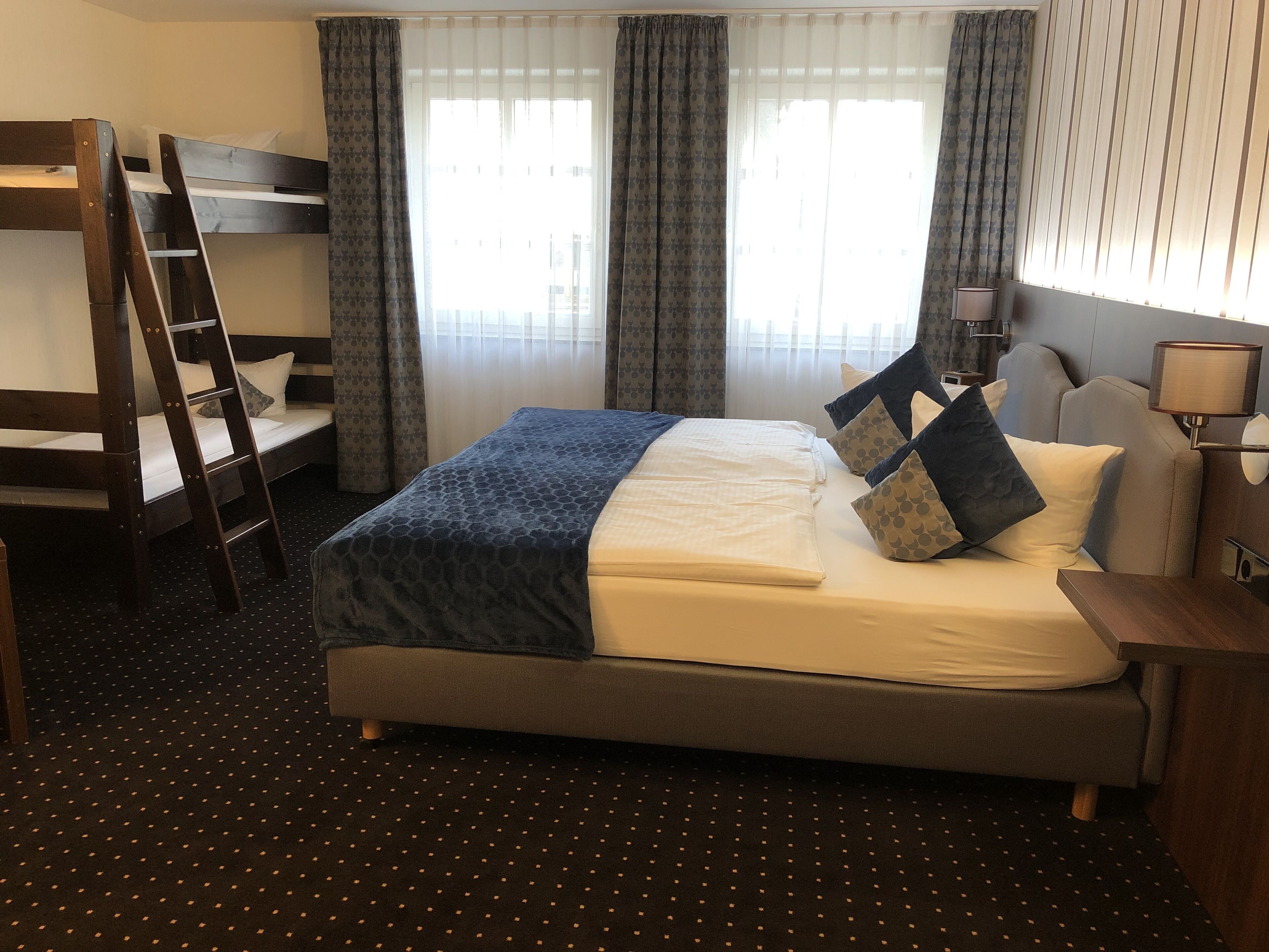 Quarto de hotel com cama beliche, escrivaninha, luminária e janela com cortinas.