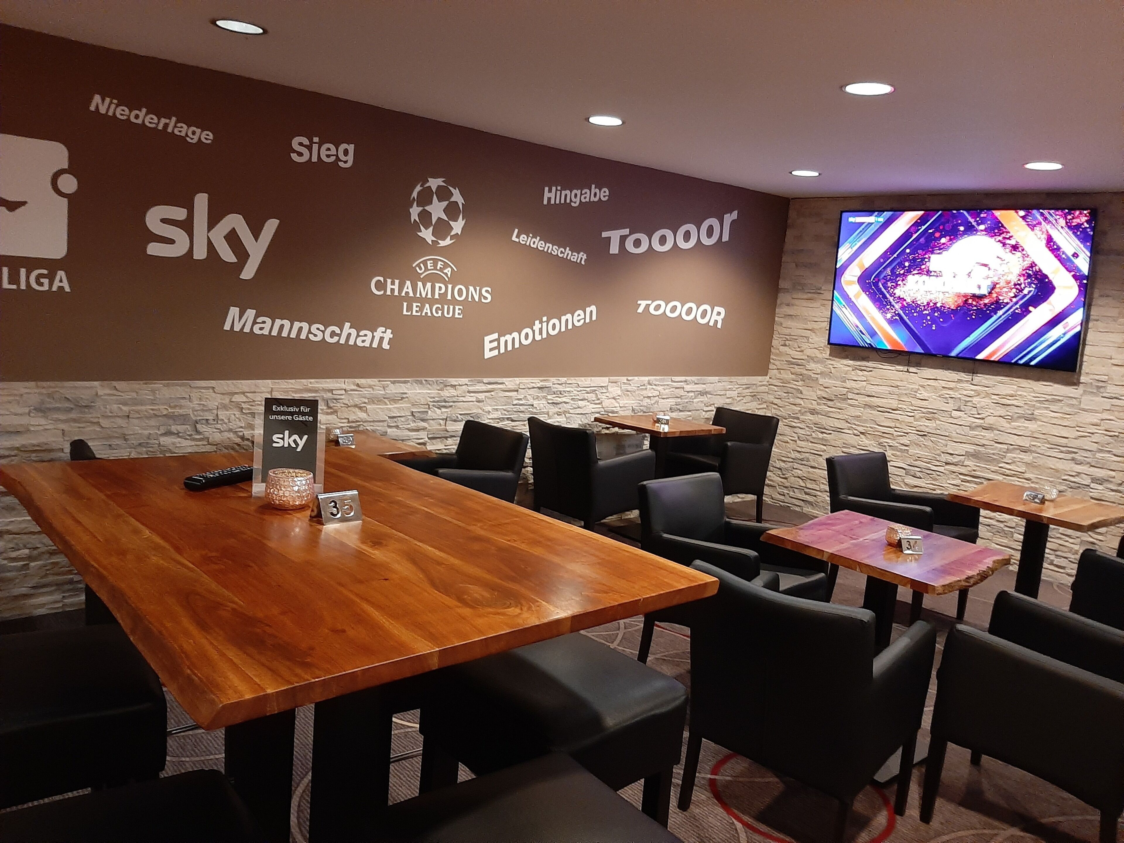 A modern lounge area with wooden tables and black chairs, a large screen displaying a sports event, and a wall with various sponsor logos.