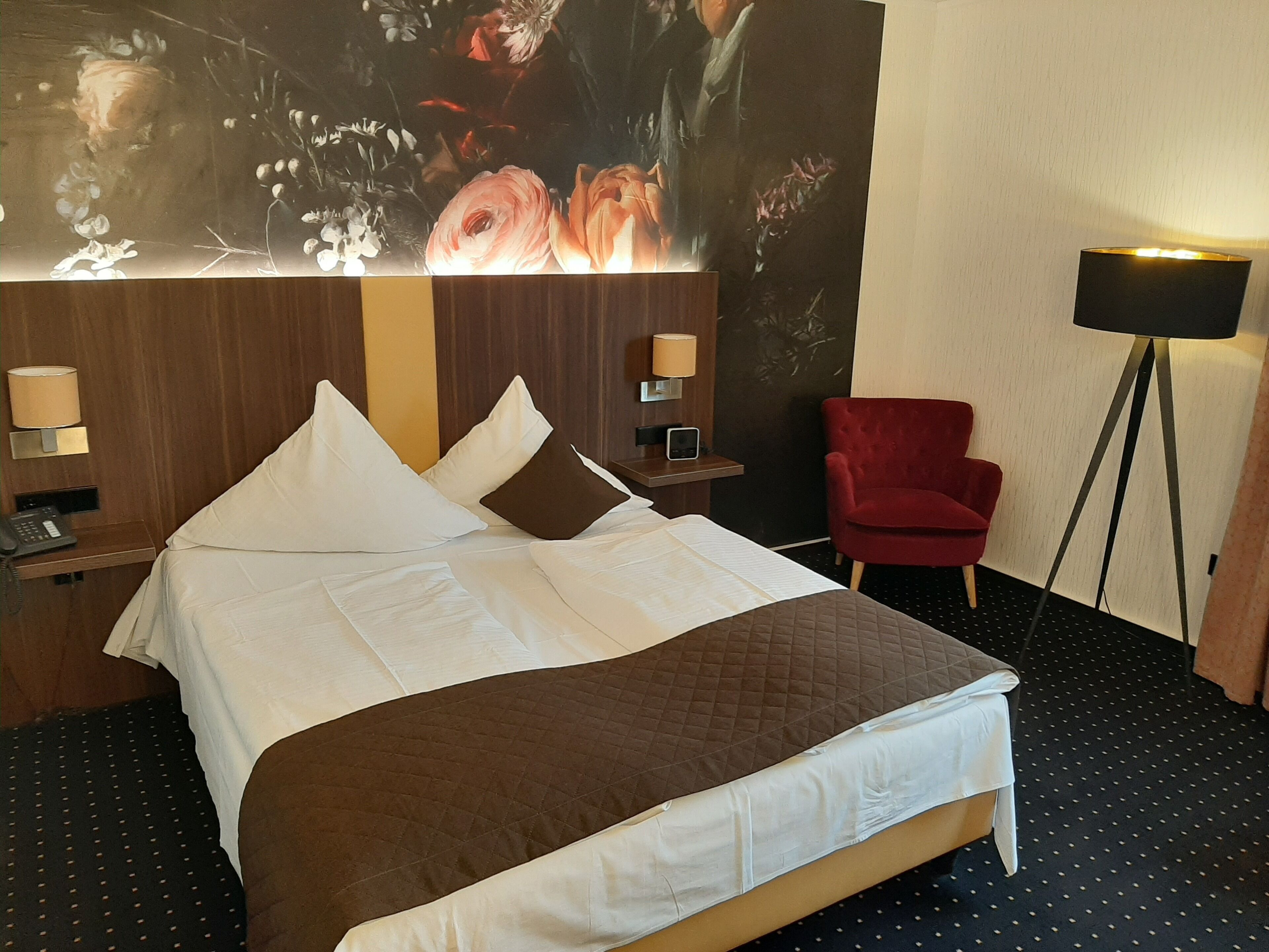 Una camera d'albergo con un letto, una poltrona rossa, una lampada da terra e un telefono fissato al muro.