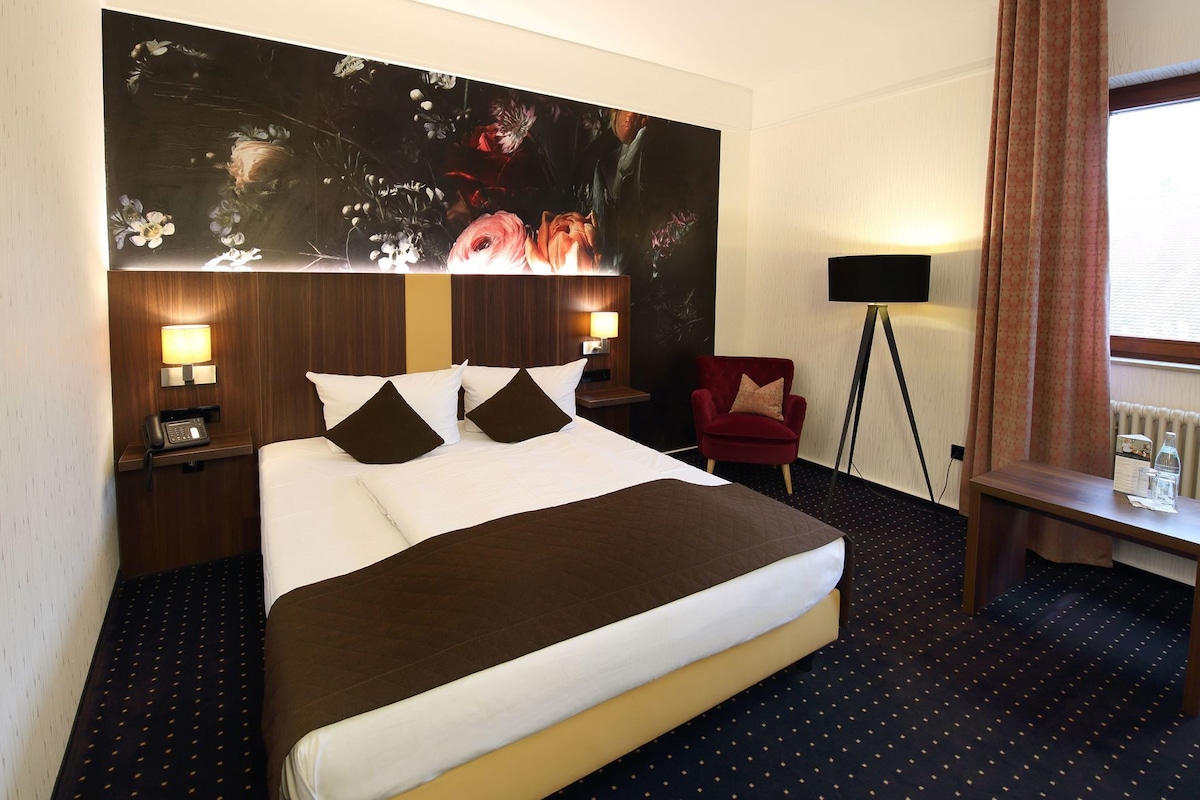 A hotel room with a large bed, two bedside lamps, a red chair, a desk with a lamp, and a large floral artwork on the wall.