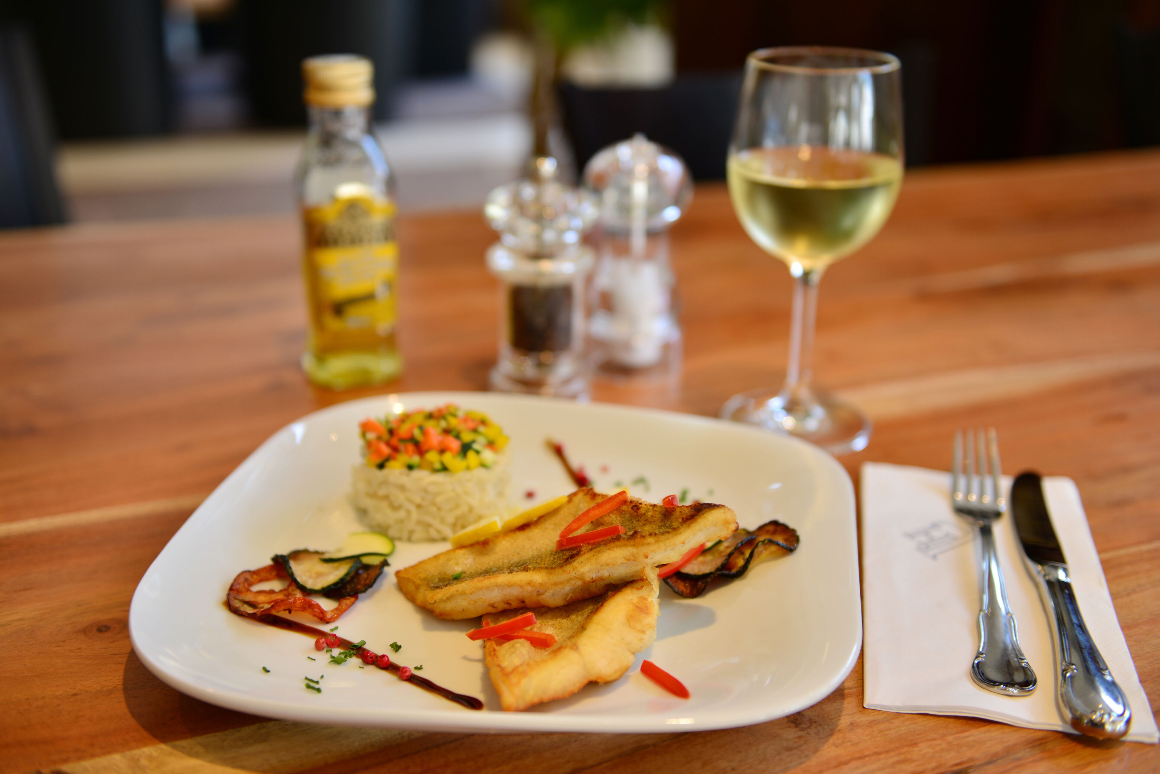 Un plato de comida con arroz, verduras y alimentos fritos, una botella de aceite, saleros y pimenteros, y una copa de vino blanco sobre una mesa de madera.