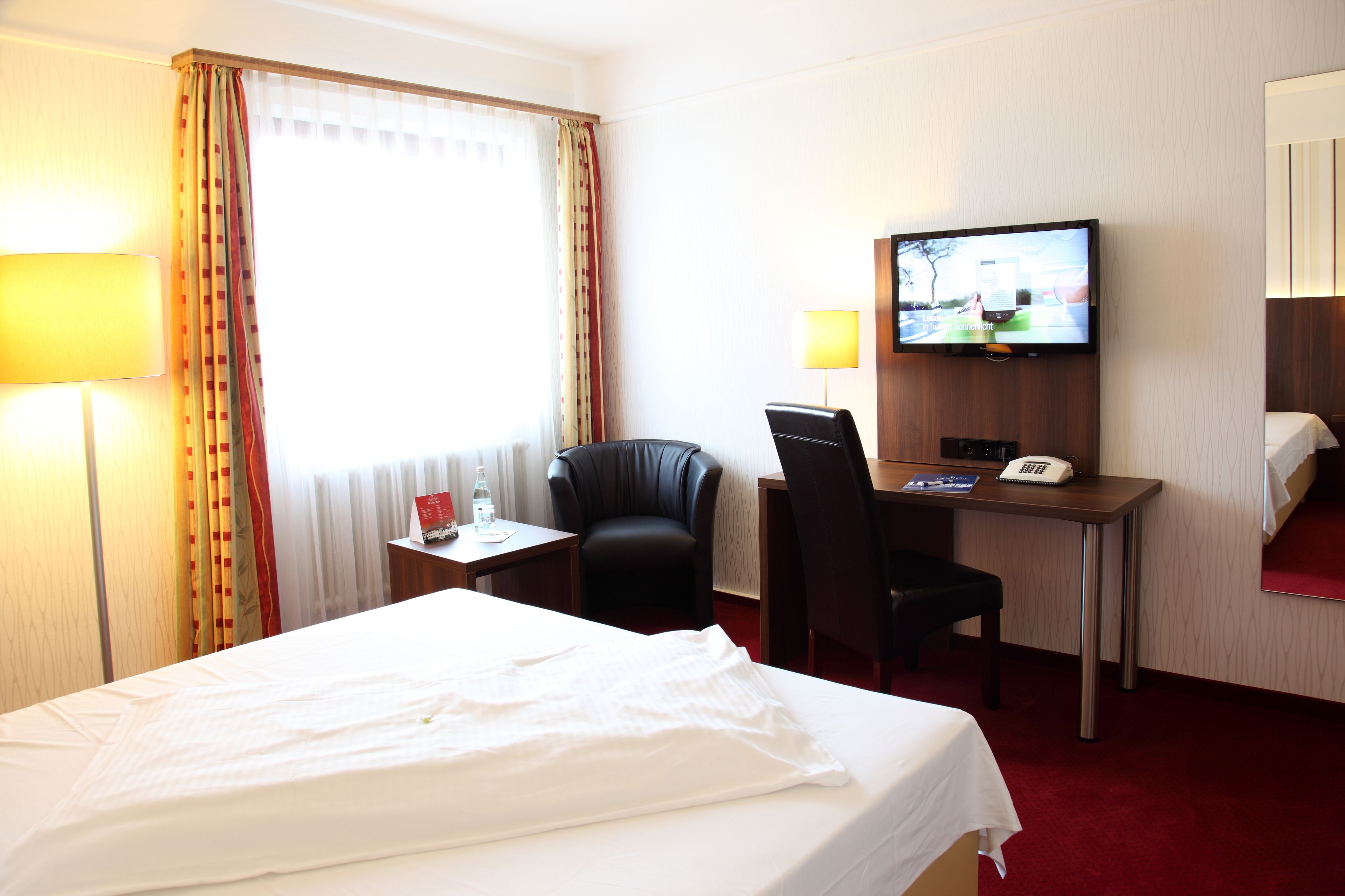 A hotel room with a bed, a desk with a chair, a television, and a small table with a lamp.
