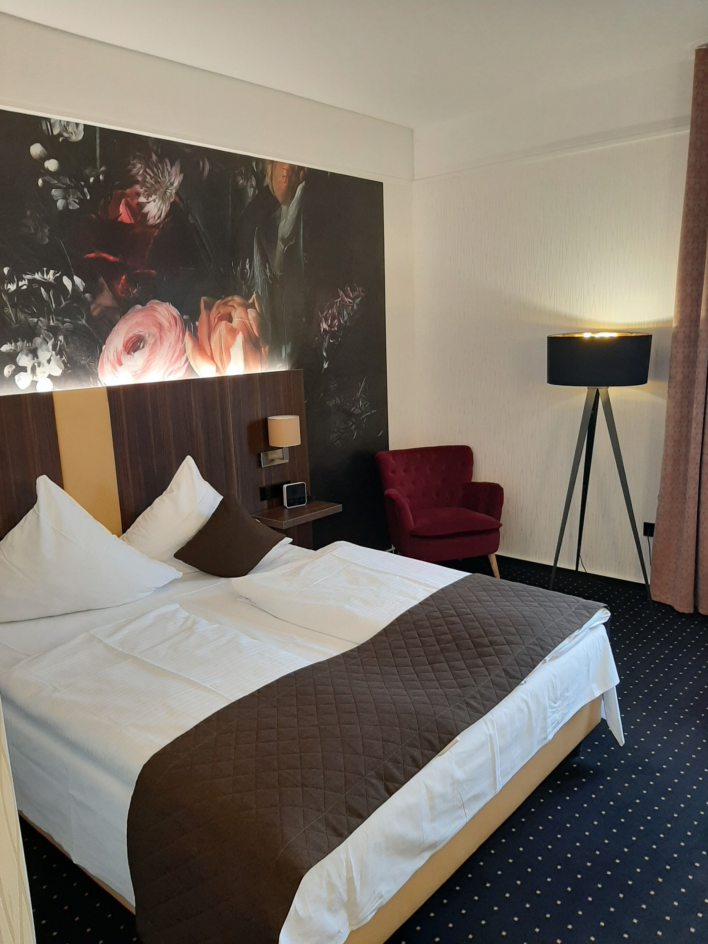 A hotel room with a large bed, a red armchair, a floor lamp, and a wall-mounted TV.