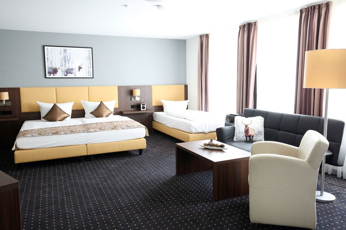 A hotel room with a large bed, a smaller bed, a sofa, a coffee table, and a lamp.