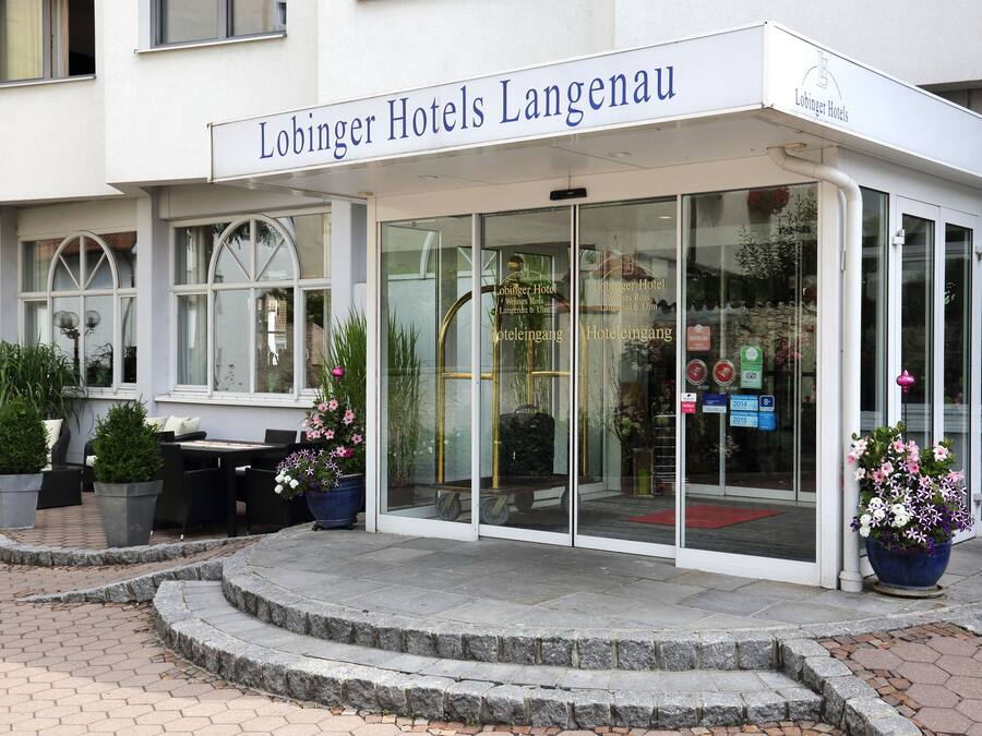 The entrance of Lohner Hotels Langenau with a glass door, potted plants, and outdoor seating.