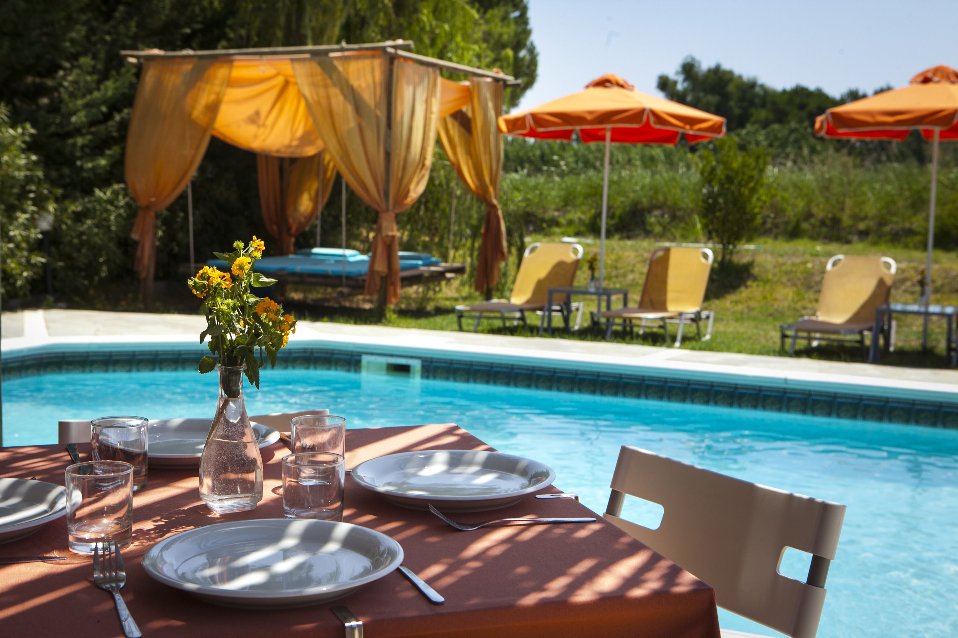 A dining table set for two by a pool with a canopy and lounge chairs in the background.