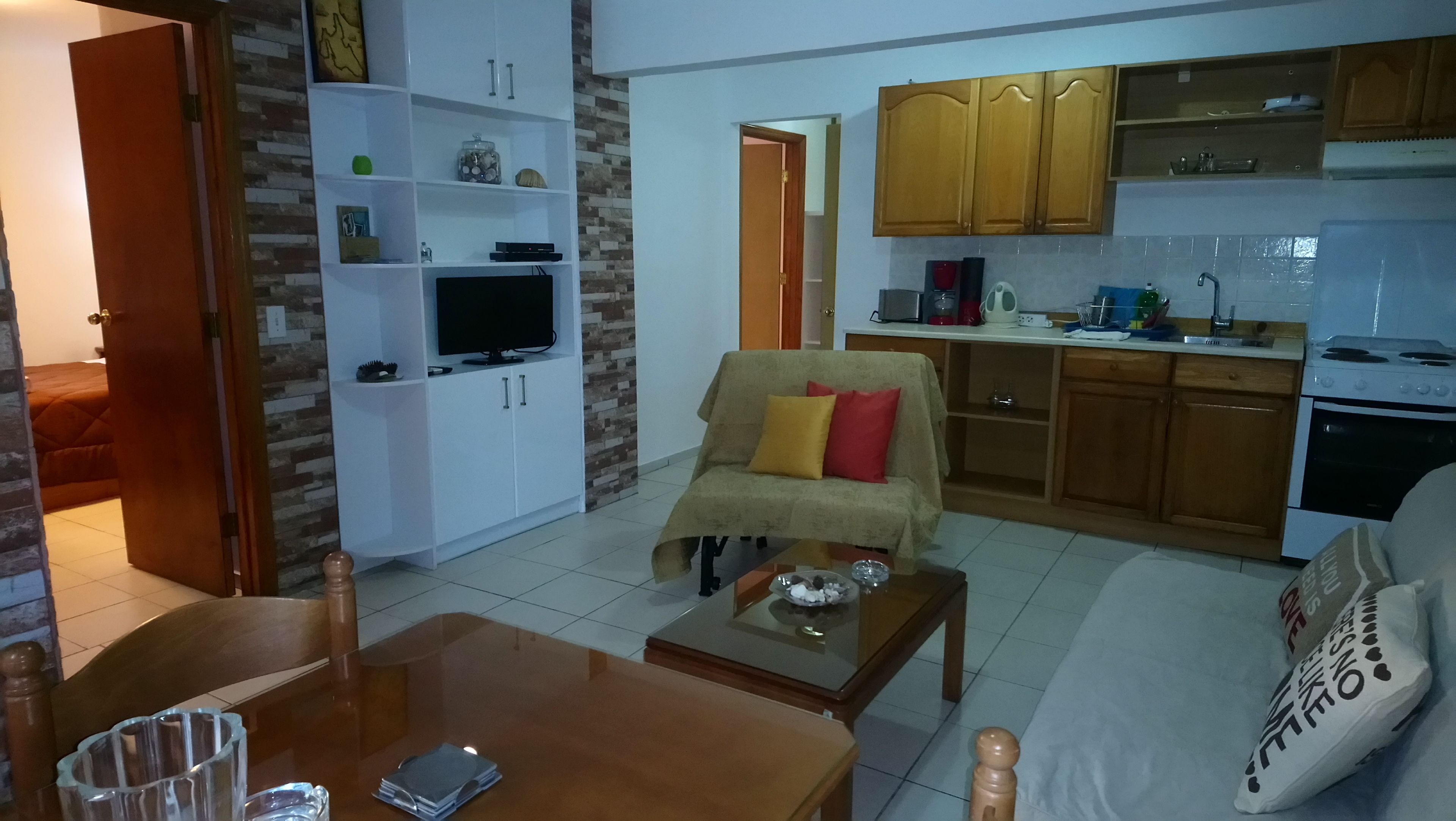 Un saló amb un sofà, taula de centre i televisor. Hi ha una zona de cuina amb armaris i electrodomèstics.