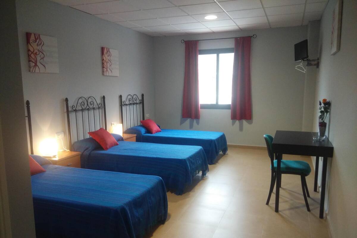 Une chambre avec quatre lits, un bureau, une chaise, une télévision et une fenêtre avec des rideaux.