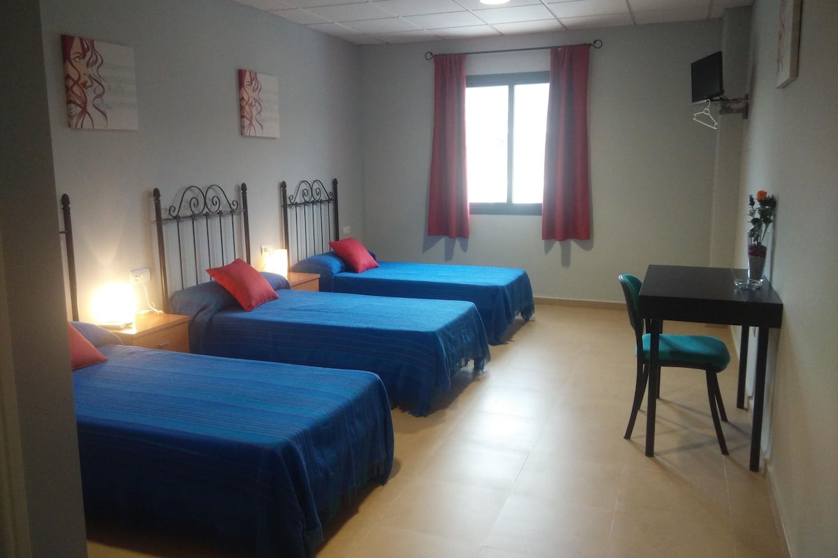 A room with four beds, a desk, a chair, a TV, and a window with curtains.