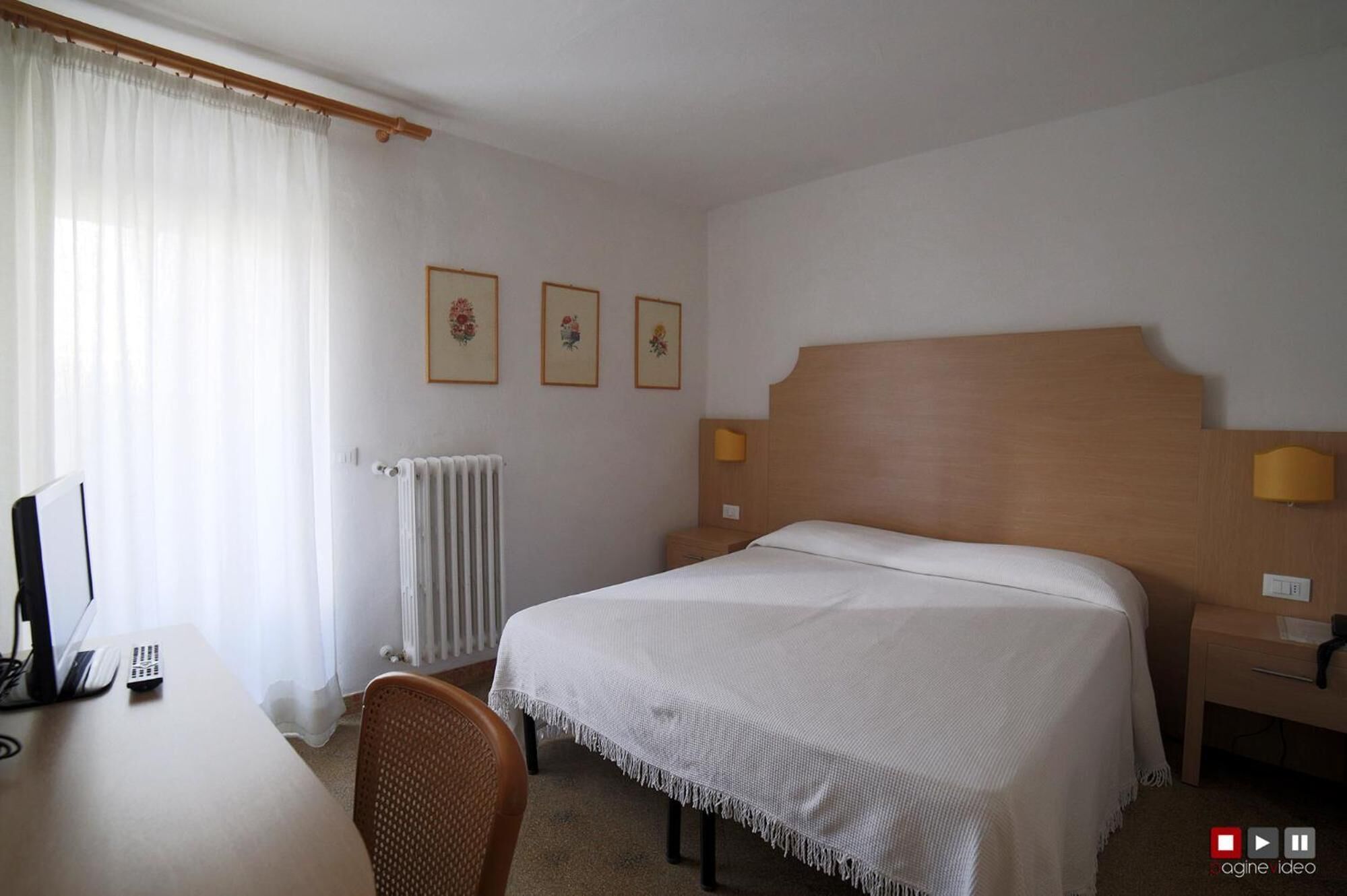 A hotel room with a bed, a desk, a chair, a radiator, and three framed pictures on the wall.