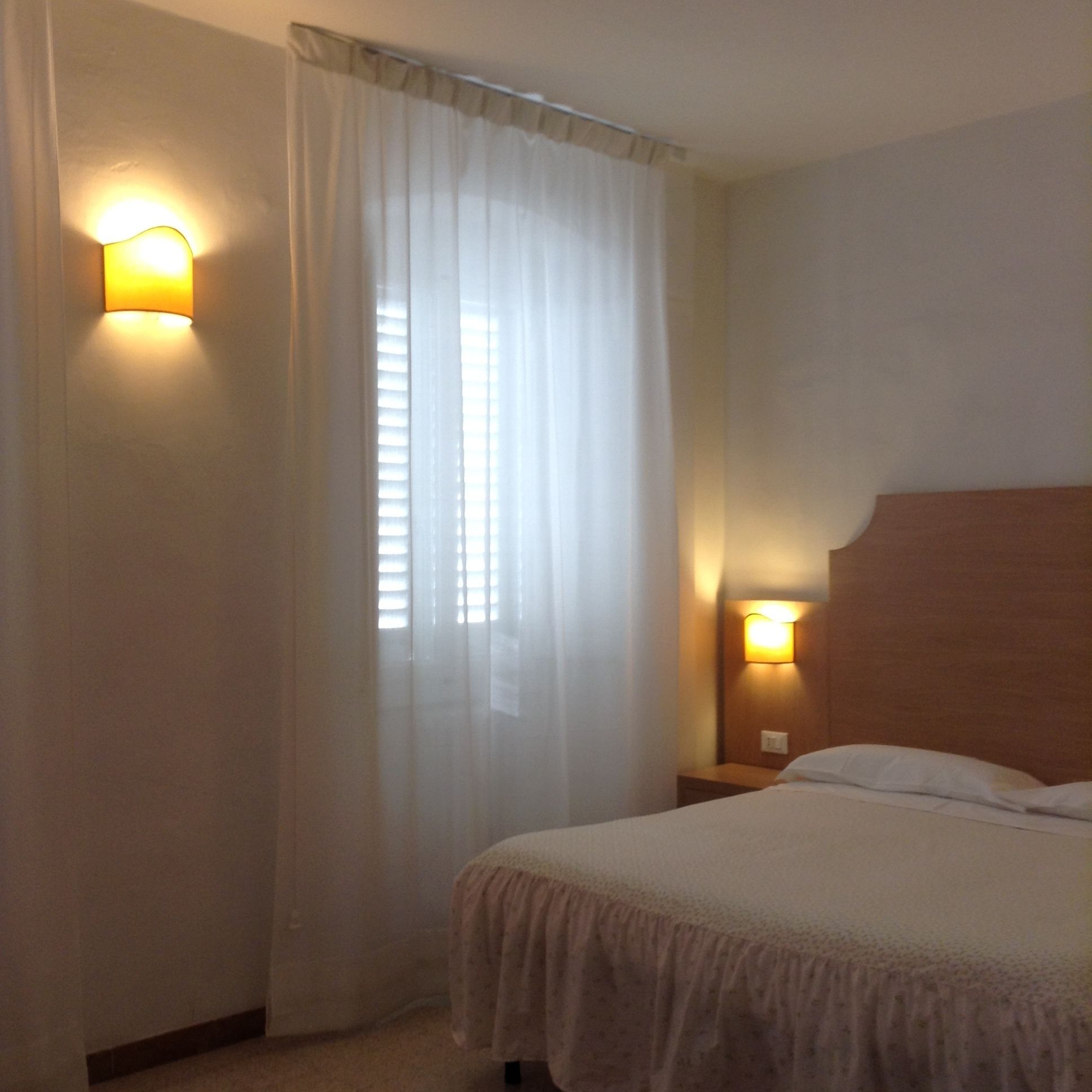 Una camera d'albergo con un letto, un comodino, una lampada a parete e una finestra con tende leggere.