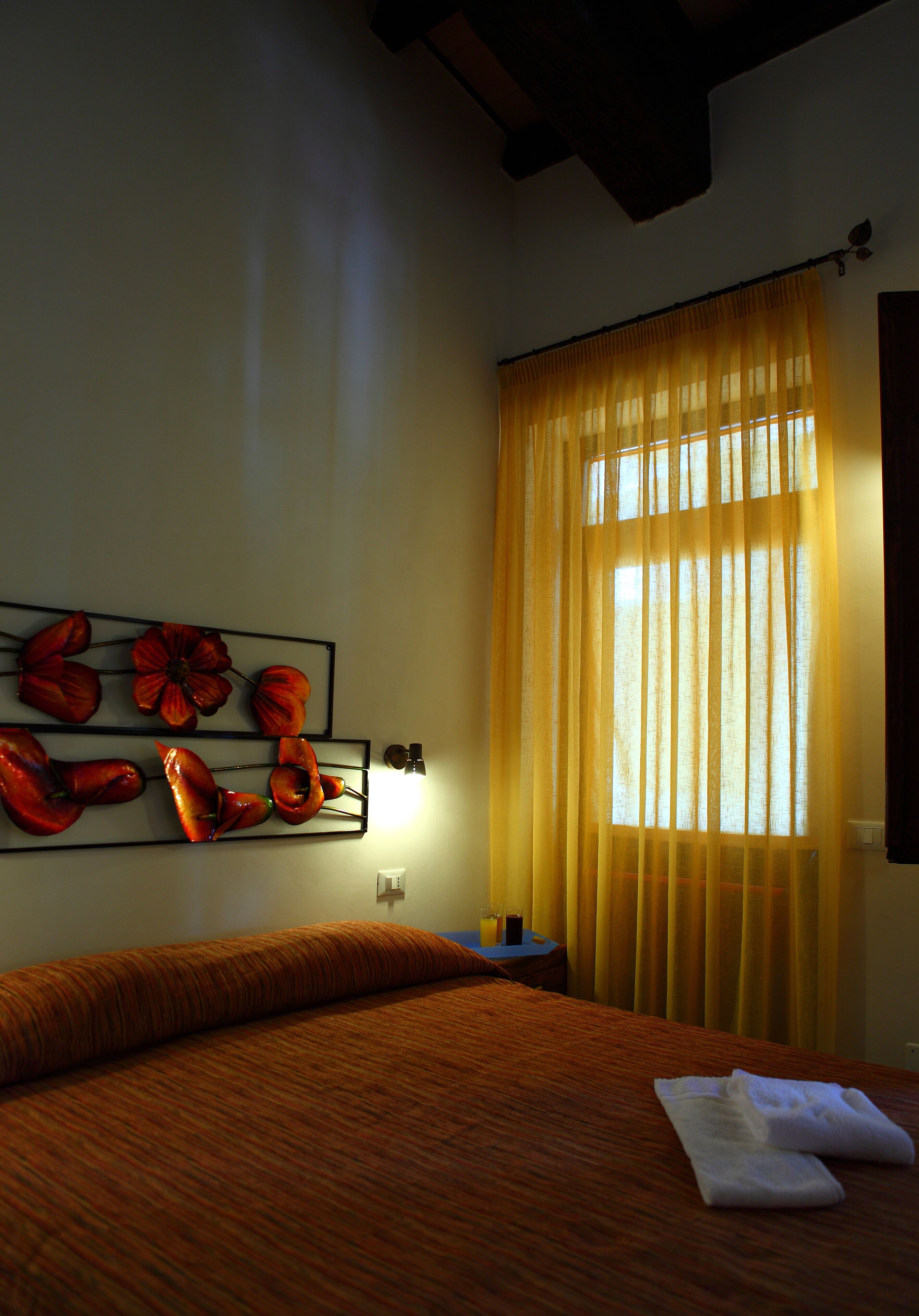 Una habitación de hotel con una cama, una obra de arte en la pared, una ventana con cortinas amarillas y una mesita de noche con lámpara.