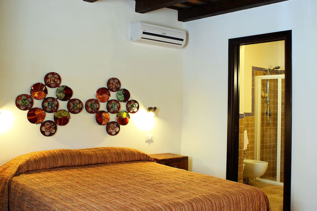 A bedroom with a bed, a wall-mounted air conditioner, a shower area, and a decorative wall piece.