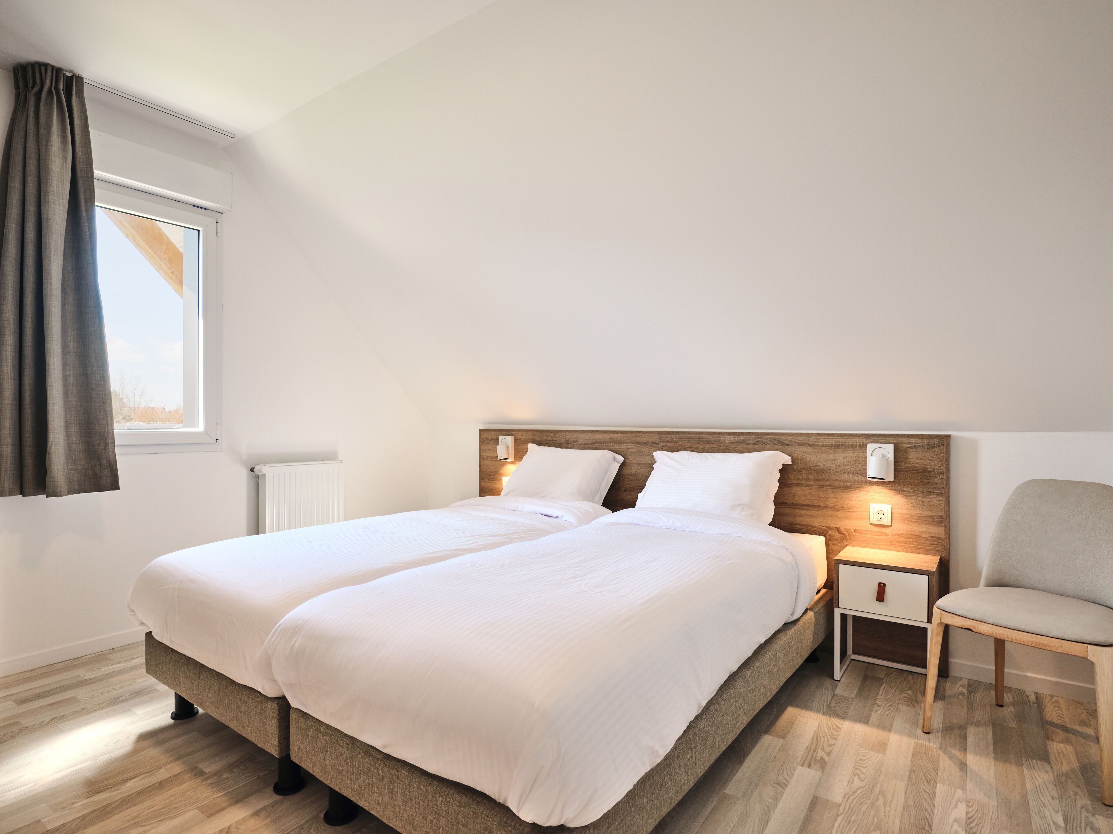 A double bed with a wooden headboard, bedside tables, a chair, and a window with curtains.