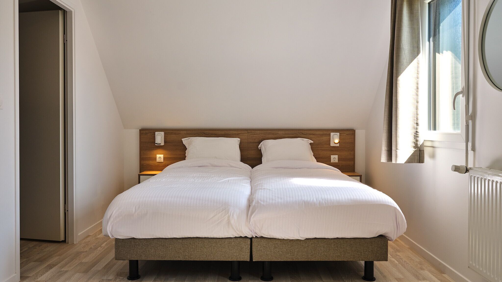 Ein Doppelbett mit Holz-Kopfteil, zwei Nachttische mit Lampen und ein Fenster mit Vorhängen.