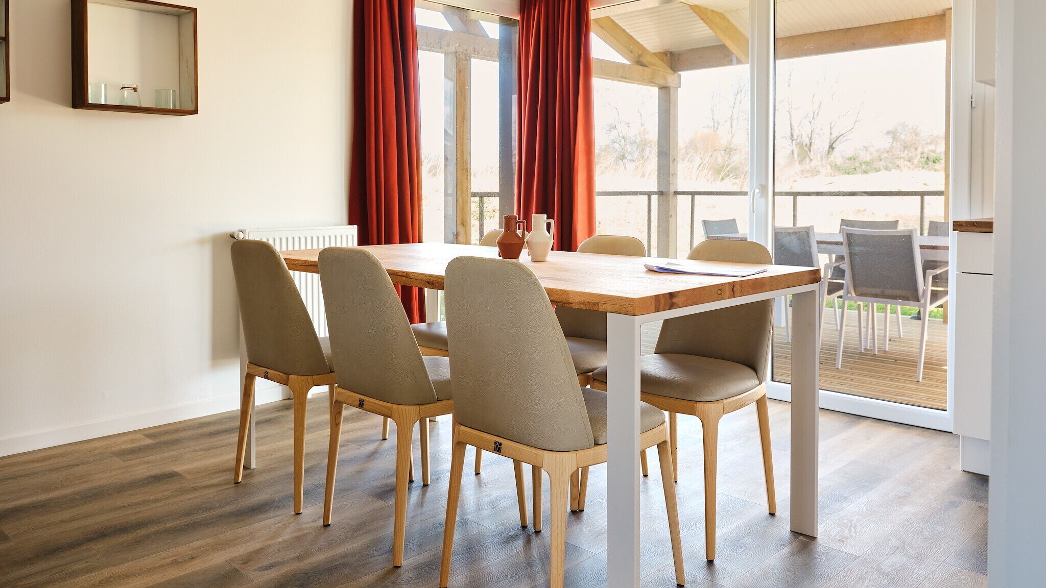 Een eethoek met een houten tafel en zes stoelen, grote ramen met rode gordijnen en uitzicht op de omgeving.