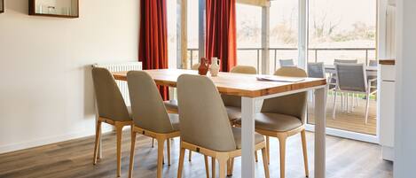 A dining area with a wooden table and chairs, large windows with red curtains, and a view of the outdoors.