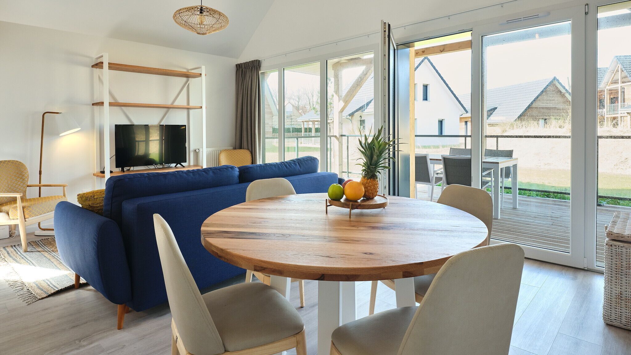 A modern living room with a blue sofa, a wooden dining table with fruit, and large windows offering a view of the outdoors.