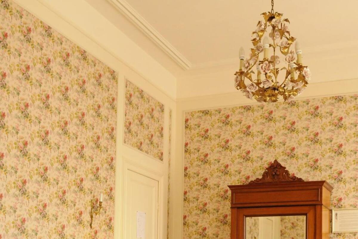 A bedroom with a large bed, floral wallpaper, a wooden wardrobe, and a chandelier.
