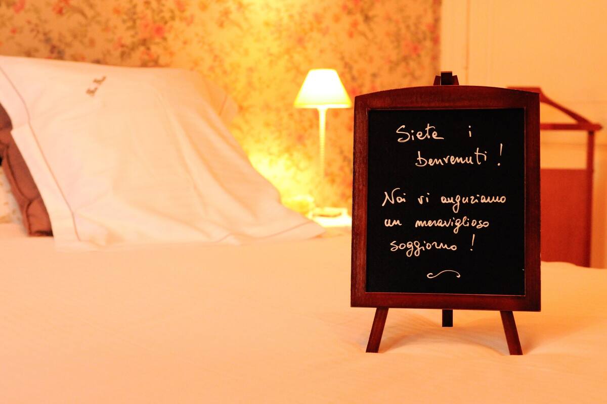 A bed with a white comforter and pillows, a bedside lamp, and a blackboard with Italian text.